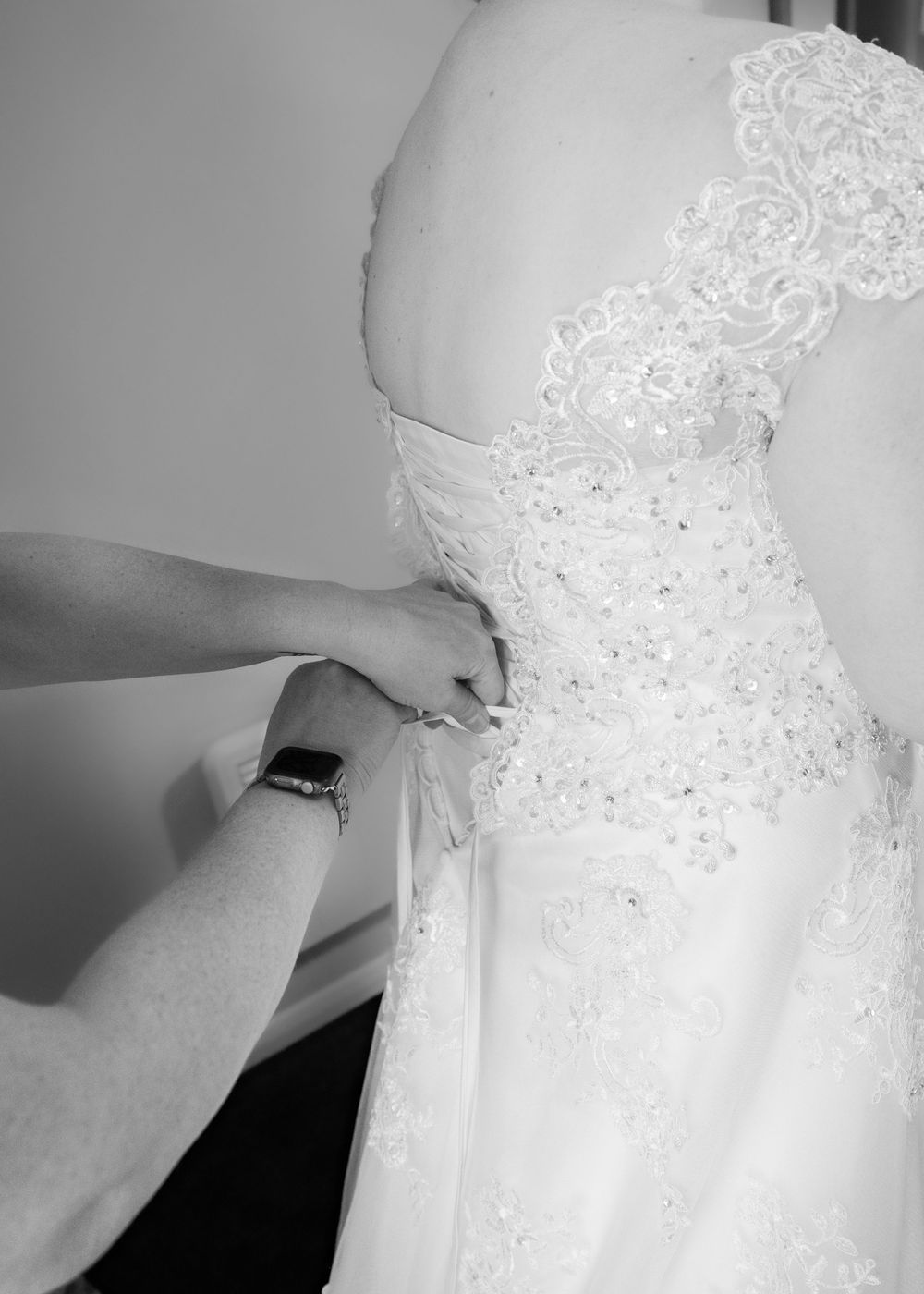 Black and white photo of a bride having her lace wedding dress fastened by a helper before the ceremony.