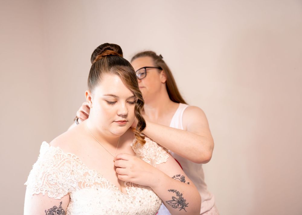 A woman in a lace wedding dress gets her hair styled by another woman in a bright studio setting.