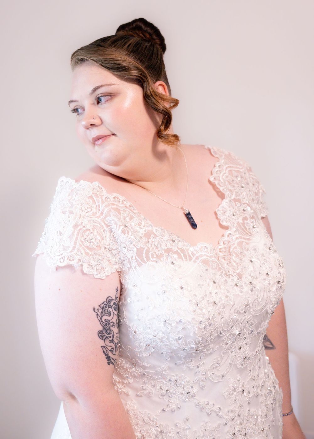 Bride in off-shoulder lace wedding gown with tattoos and crystal necklace poses elegantly.