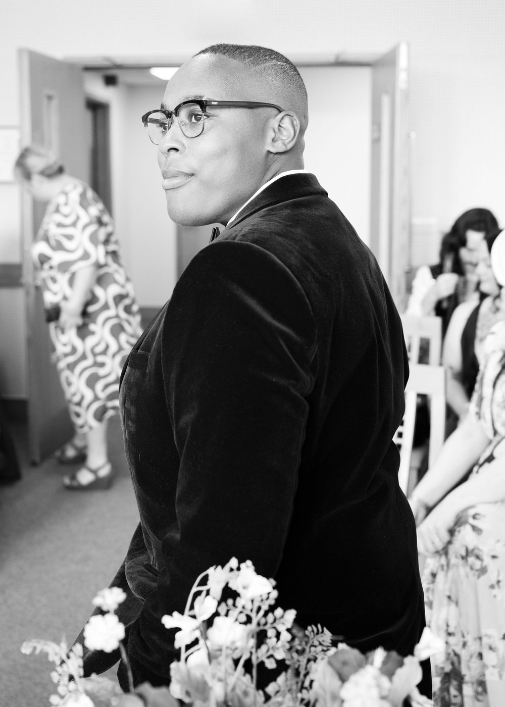 Groom in velvet blazer and glasses looks over shoulder at wedding ceremony in black and white photo.