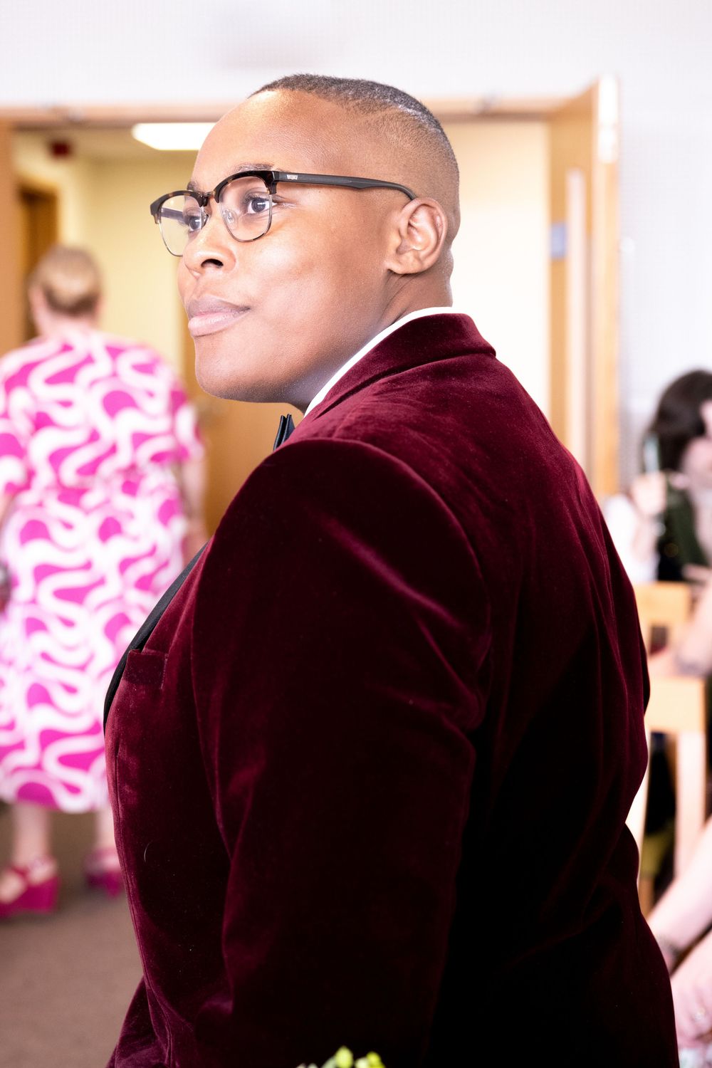 Stylish man in burgundy velvet blazer and glasses looking over shoulder in elegant indoor setting.