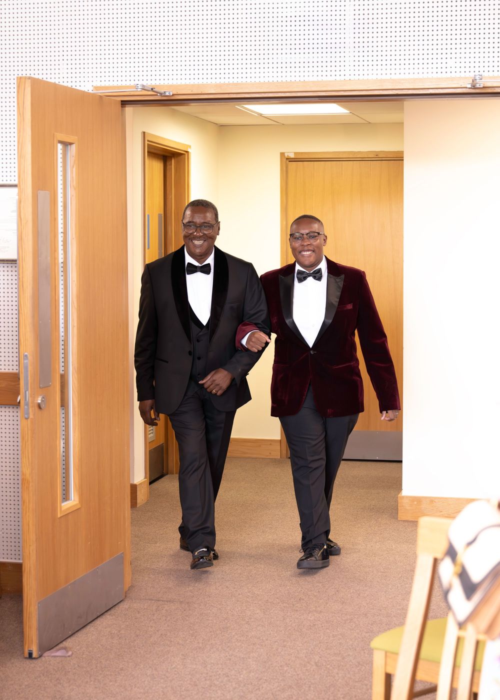 Two men in formal tuxedos smiling and walking through a doorway, one in black and one in burgundy velvet jacket.