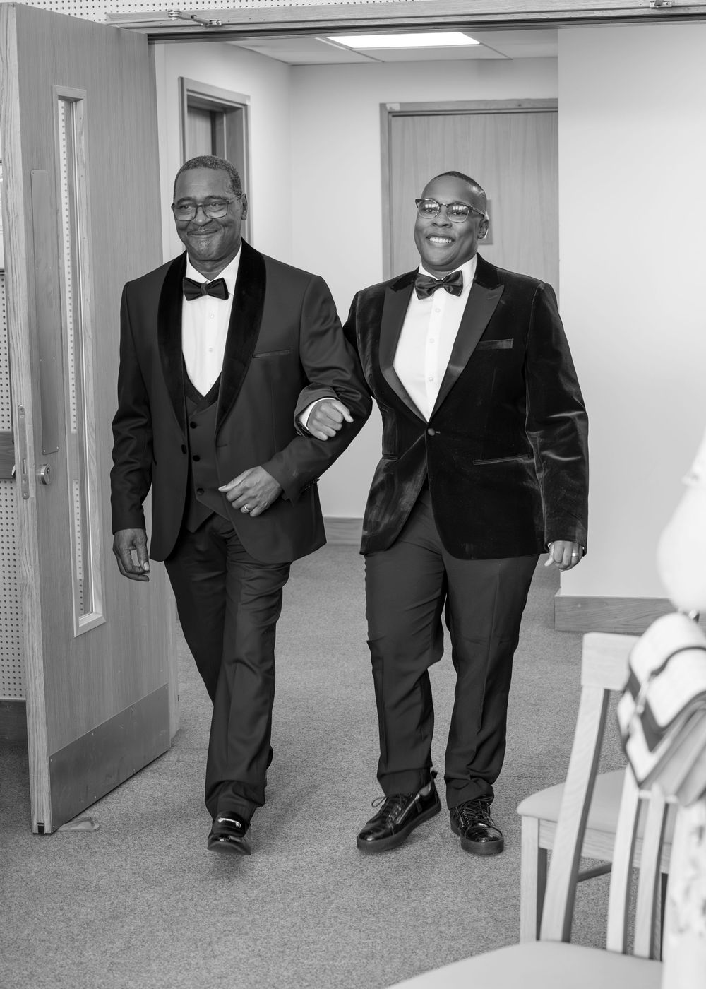 Two men in tuxedos with bow ties walk together smiling in a hallway, black and white photo.