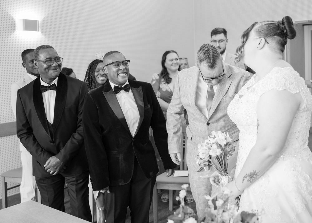 Black and white wedding photo of guests smiling and laughing with groom and bride at ceremony altar.