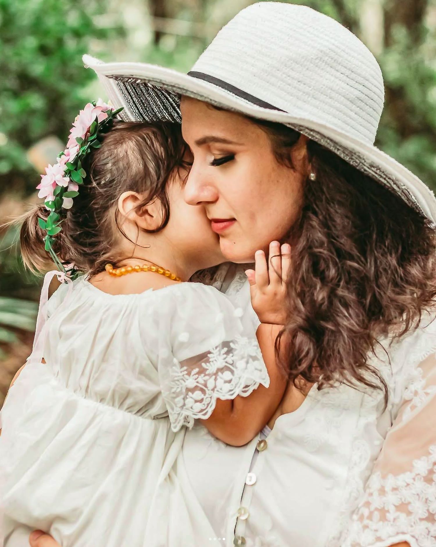 A young girl with flower crown kisses a woman in white hat and dress outdoors at Delray Oaks Natural Area.