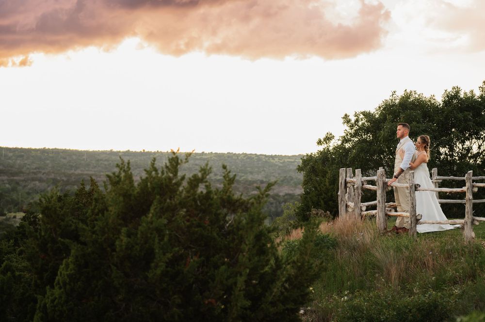 Couple in wedding attire standing by rustic wooden fence overlooking scenic Texas Hill Country landscape at sunset.