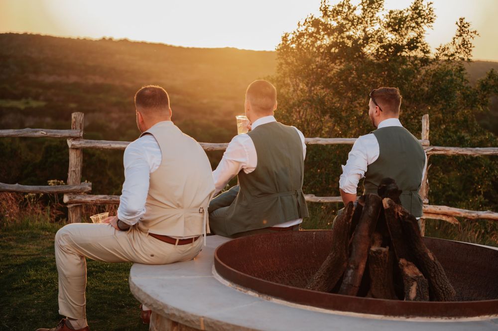 Three groomsmen in vests sit by a fire pit at sunset, enjoying drinks with a scenic countryside view.