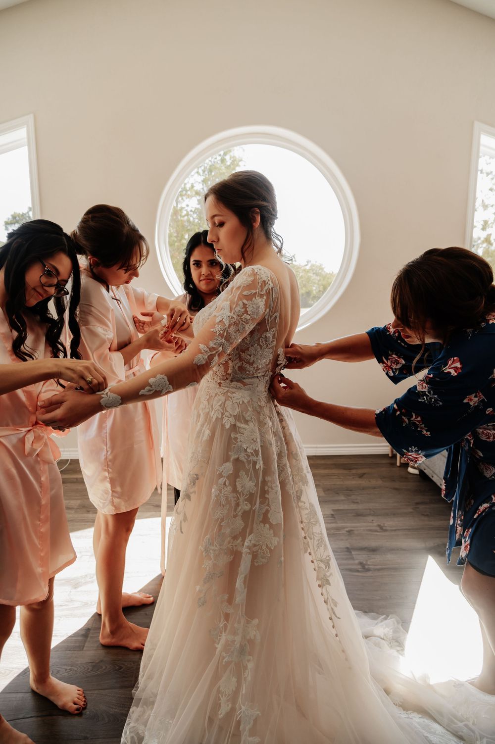 Bridesmaids in pink robes helping bride into lace wedding gown near round window in bright room.