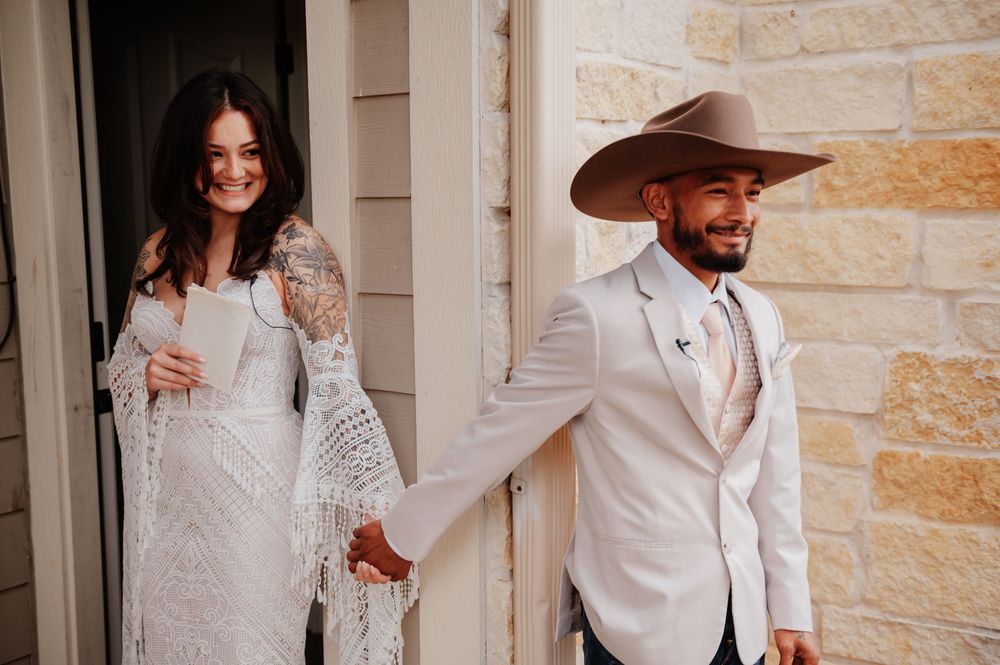 Bride in lace boho dress and groom in white suit with tan cowboy hat hold hands at rustic wedding venue.