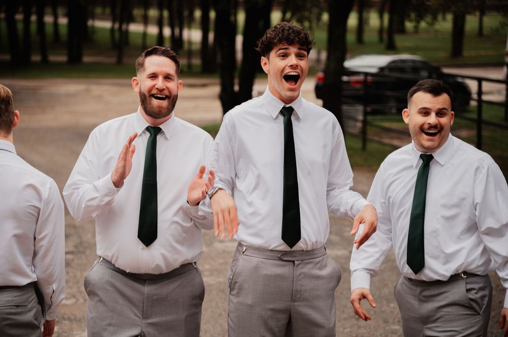 Three groomsmen in white shirts and green ties laughing and celebrating outdoors at a wedding.