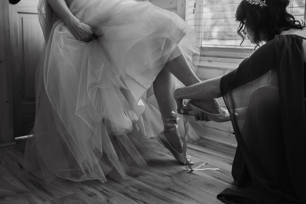 Black and white photo of bridesmaid kneeling to fasten bride's wedding shoes while holding up elegant bridal gown.
