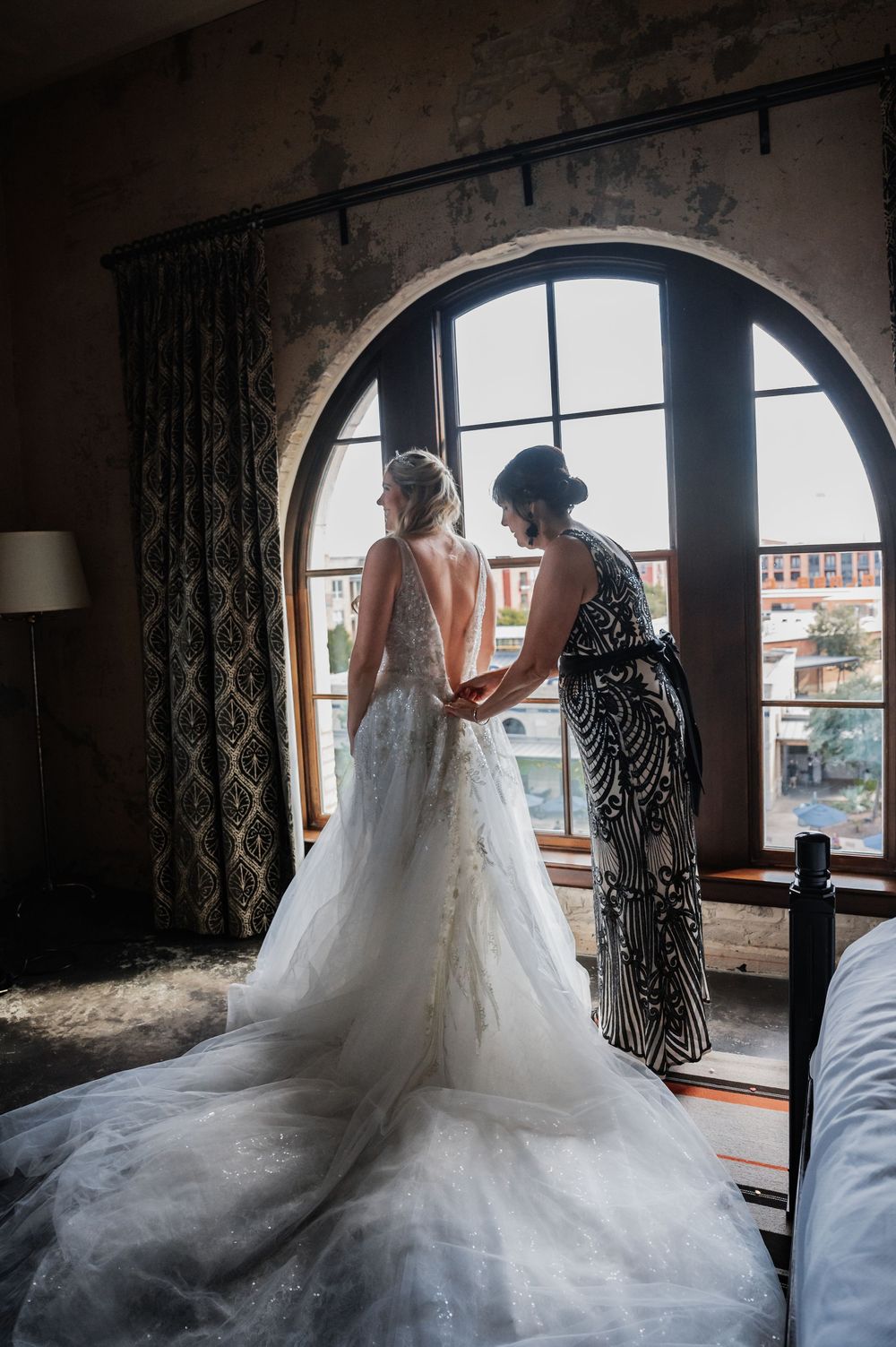 Bride in lace gown with long train stands by arched window as bridesmaid helps fasten her dress.