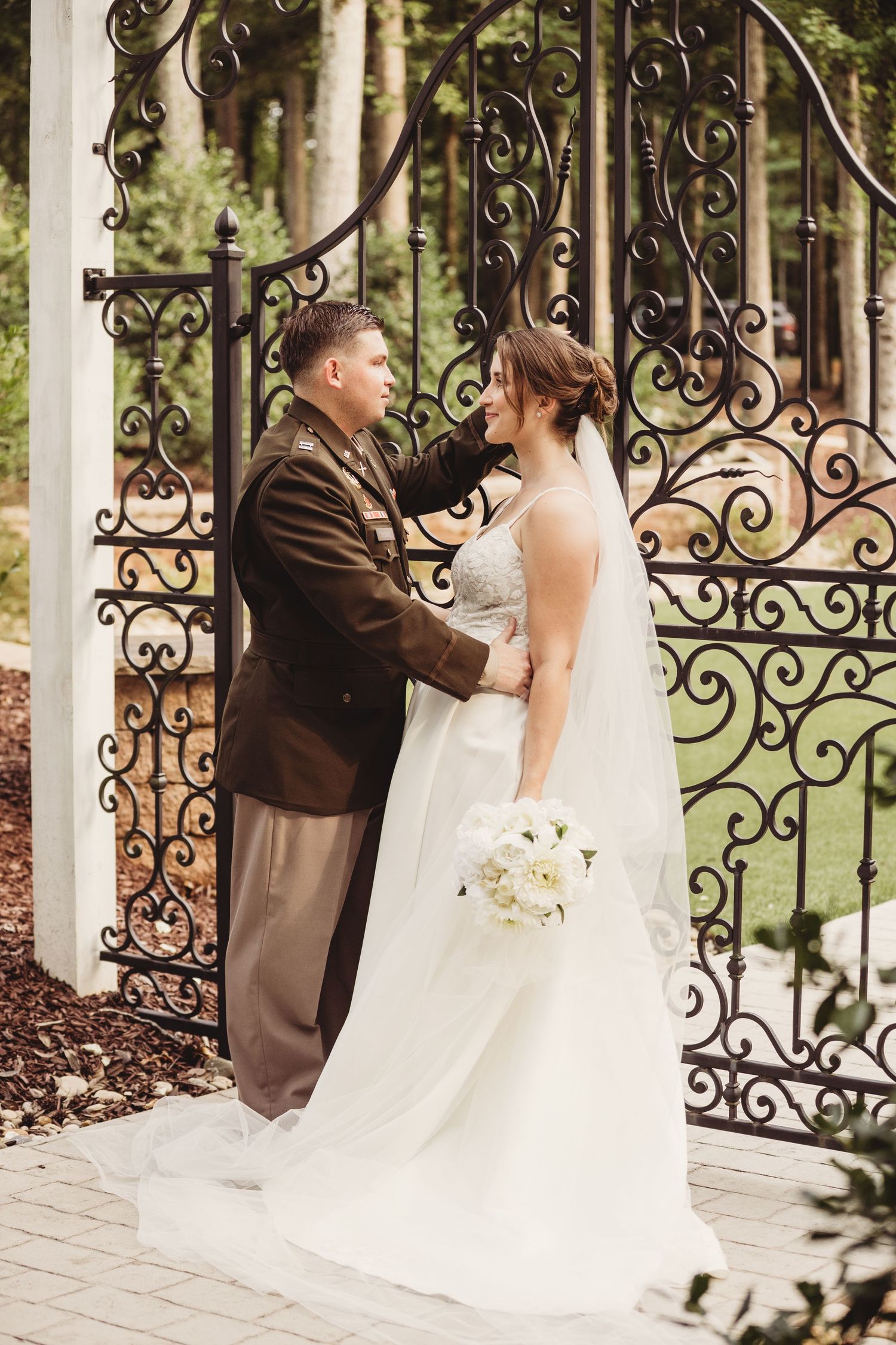 Military groom in dress uniform holds hands with bride in white gown before ornate iron garden gates.