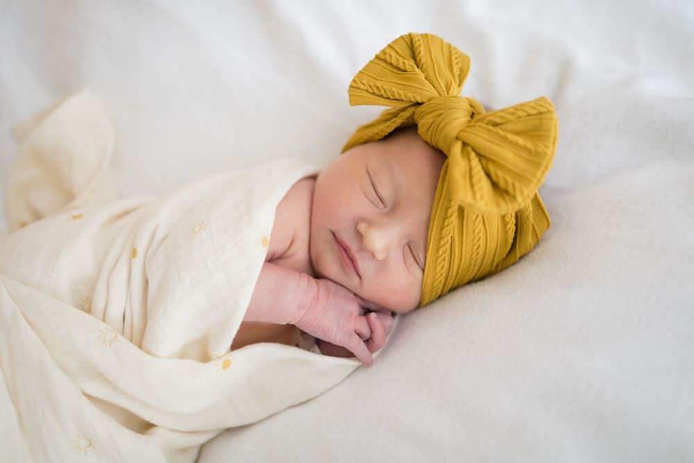 Sleeping newborn baby swaddled in white wrap wearing a mustard yellow knit bow headband. Images at Fort Sanders Medical Center in Knoxville.