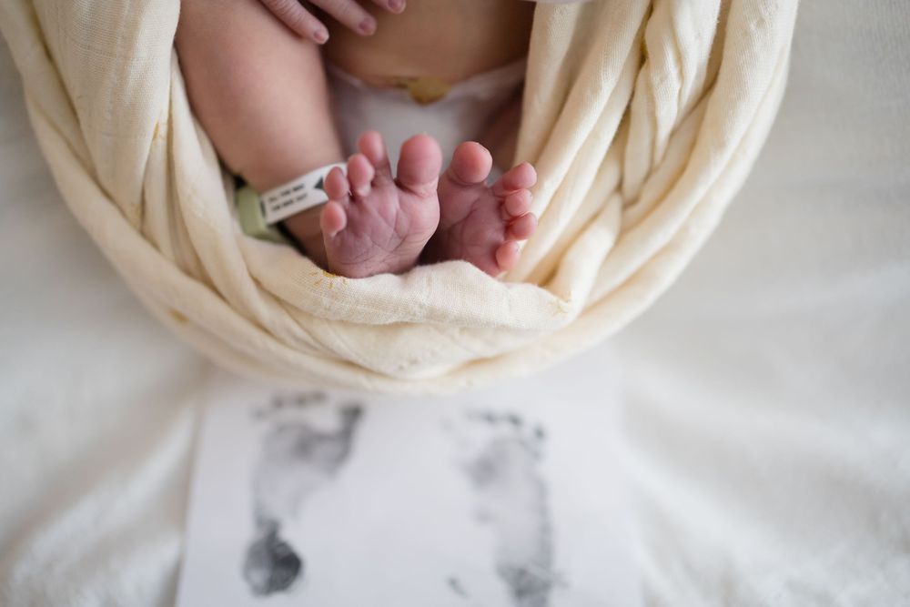 Newborn baby feet wrapped in cream swaddle blanket with hospital ID bracelet and footprint card below. Images at Fort Sanders Medical Center