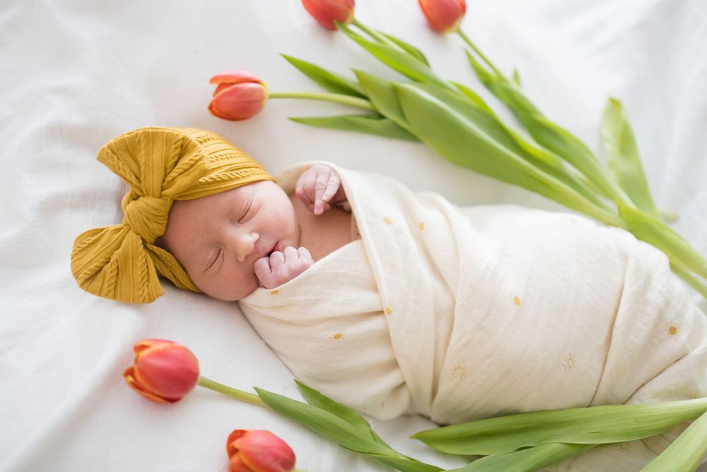 Sleeping newborn baby swaddled in cream wrap with yellow bow headband, surrounded by red tulips on white background at Fort Sanders Regional