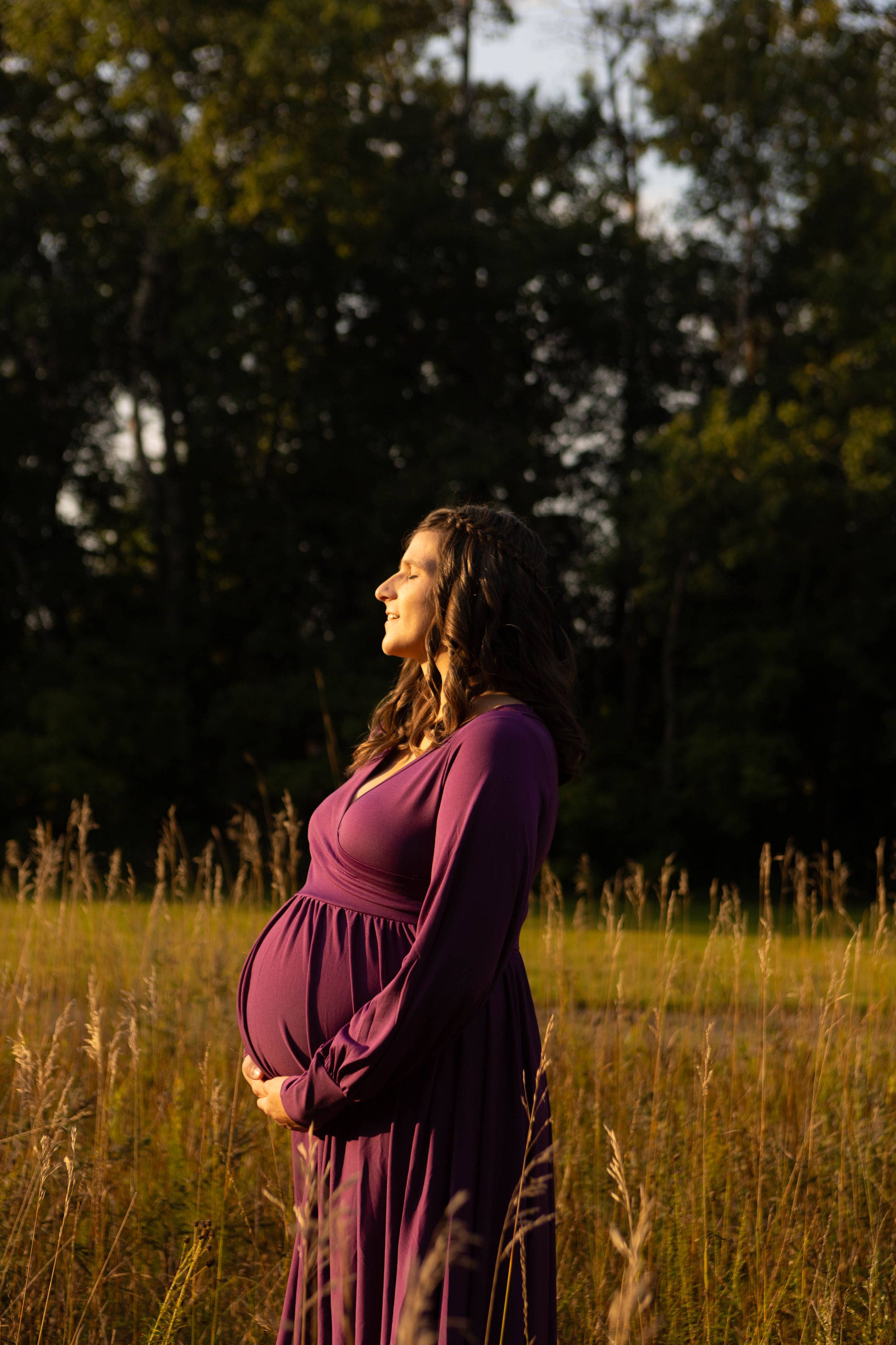 Pregnant woman in purple dress standing in golden field at sunset, cradling baby bump with trees in background.