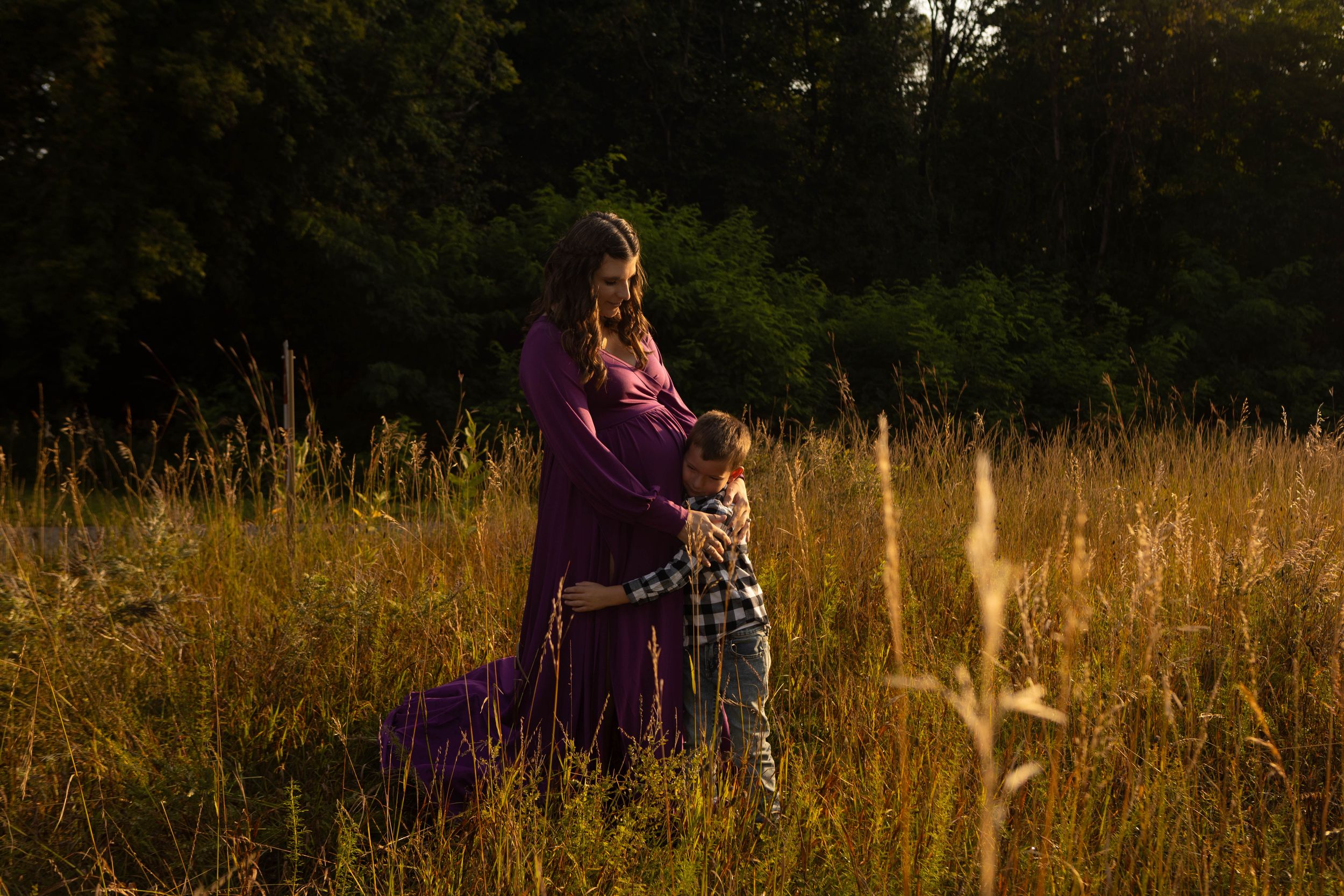 Pregnant woman in purple dress standing in golden field with young child hugging her belly at sunset.