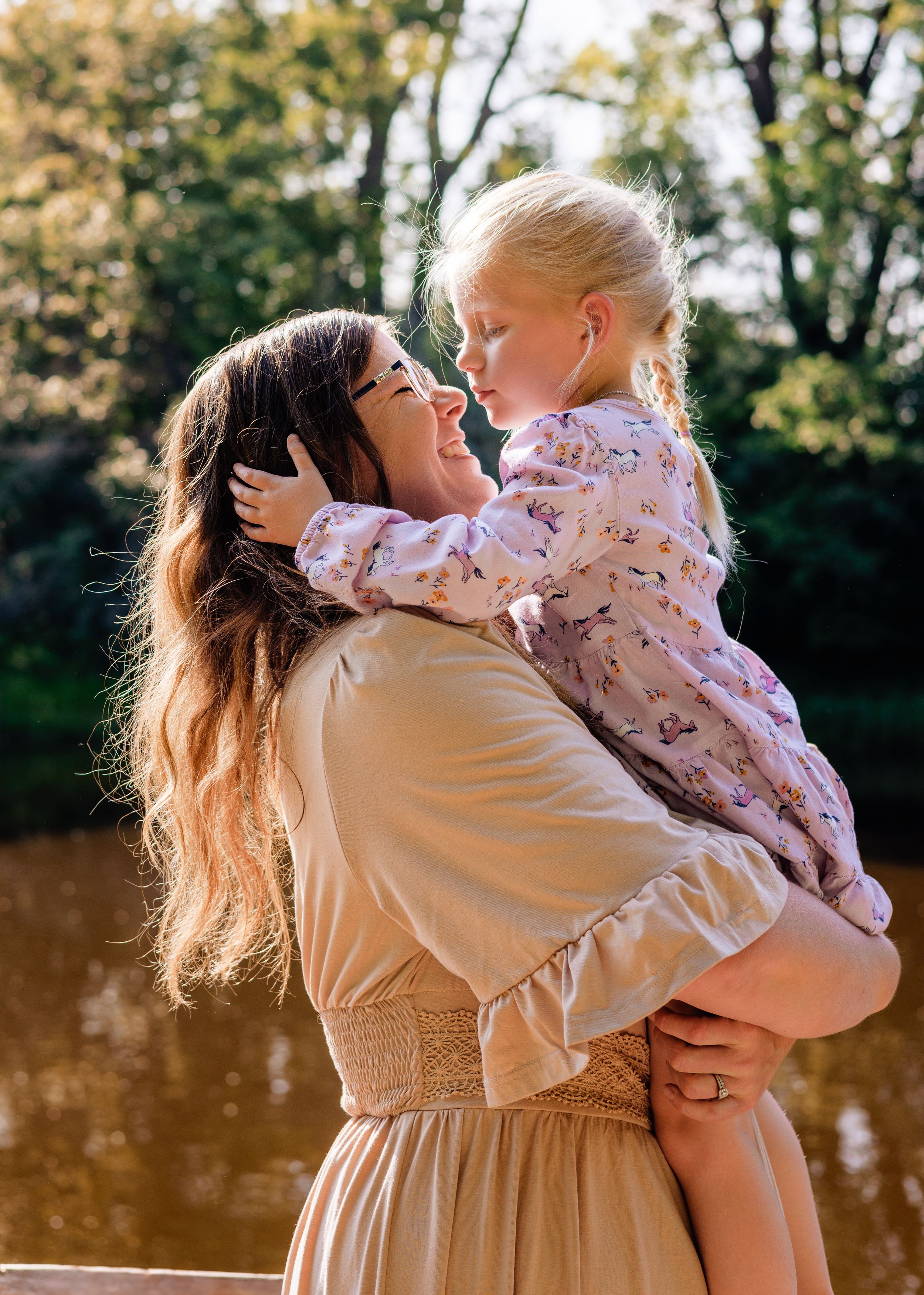 A mother holds her young blonde daughter outdoors near a sunlit pond, both smiling and sharing a tender moment.