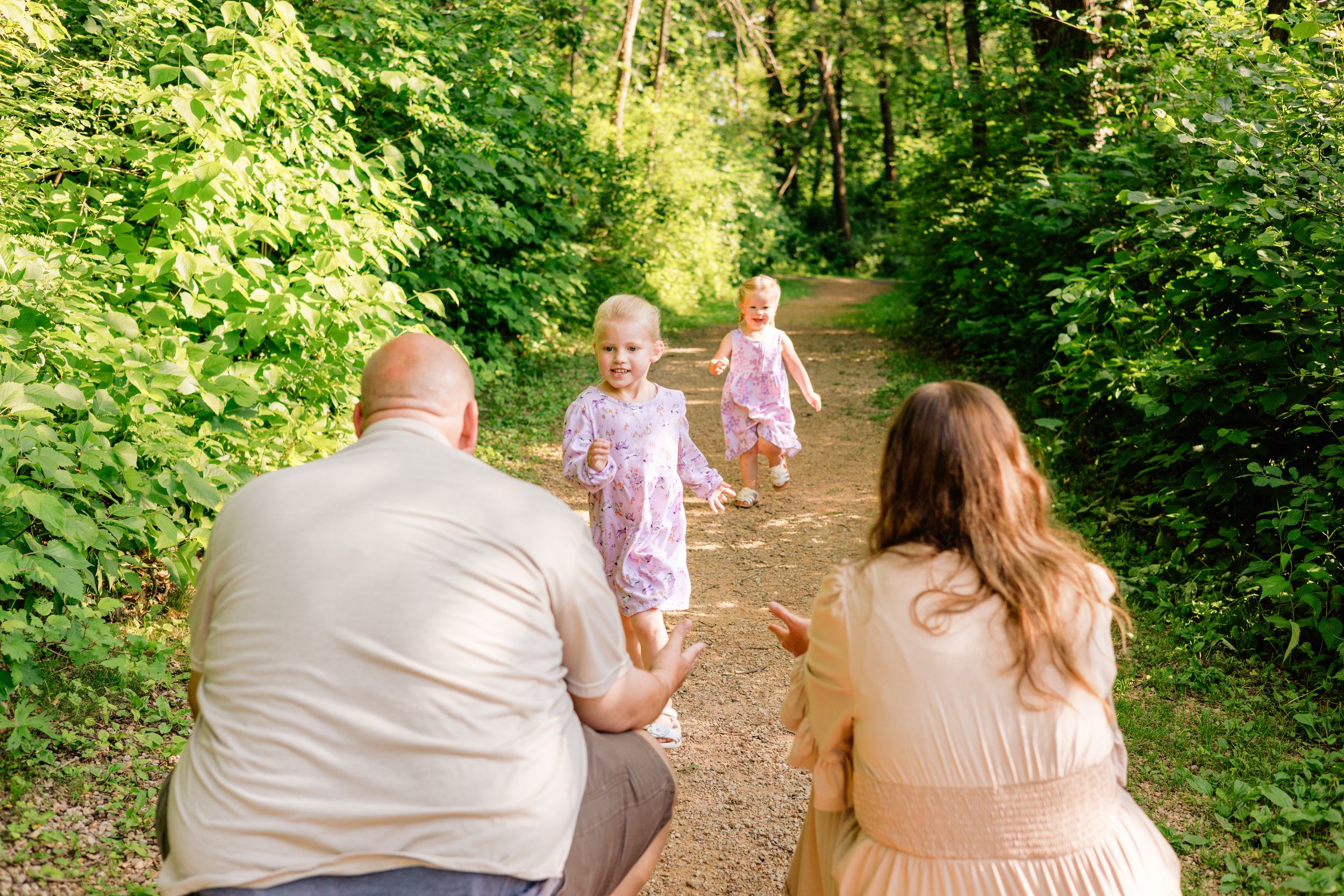 Family sitting on a forest path watching two young girls in purple dresses running toward them outdoors.
