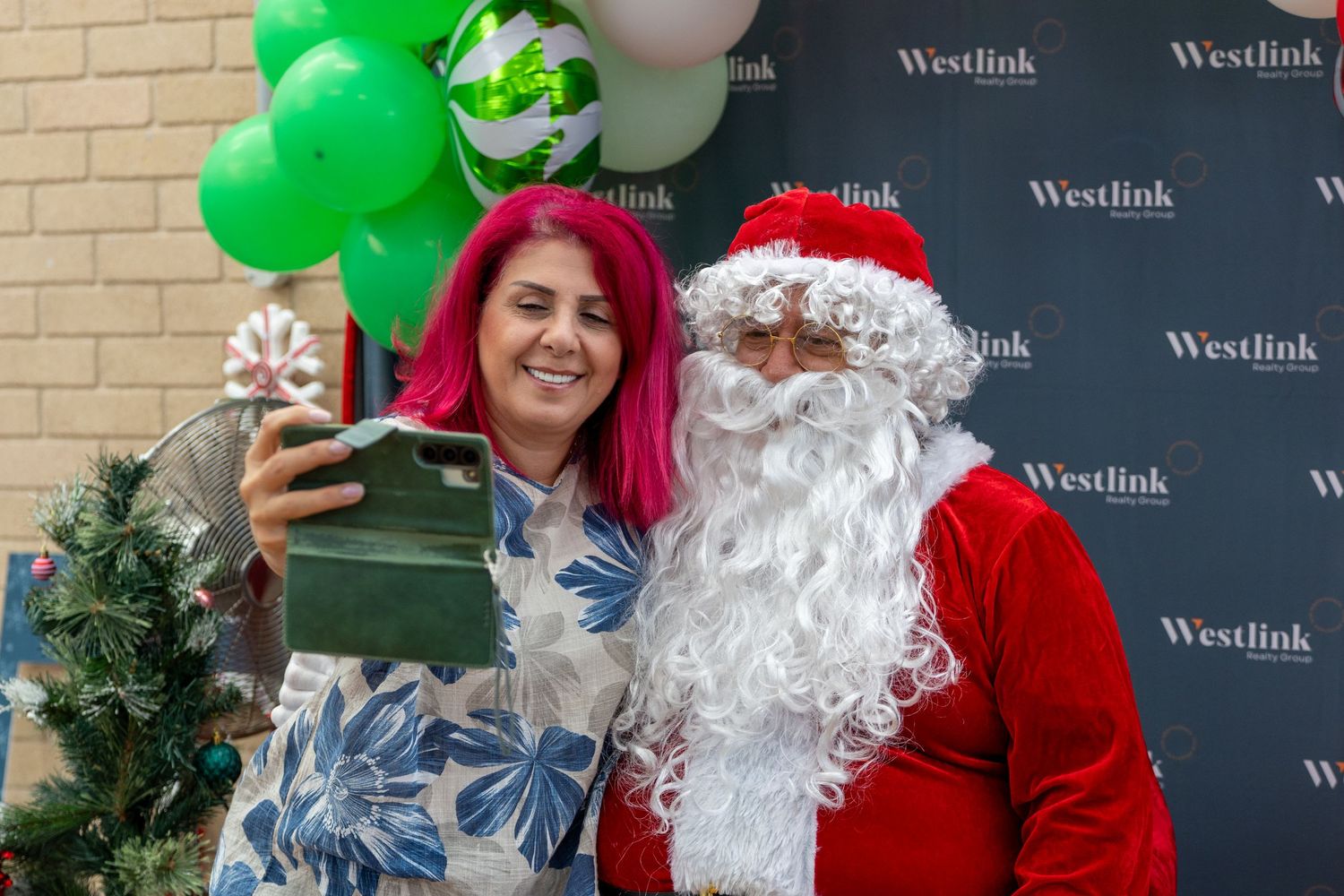 Woman with pink hair taking a selfie with Santa Claus at a Westlink Christmas event with green balloons.