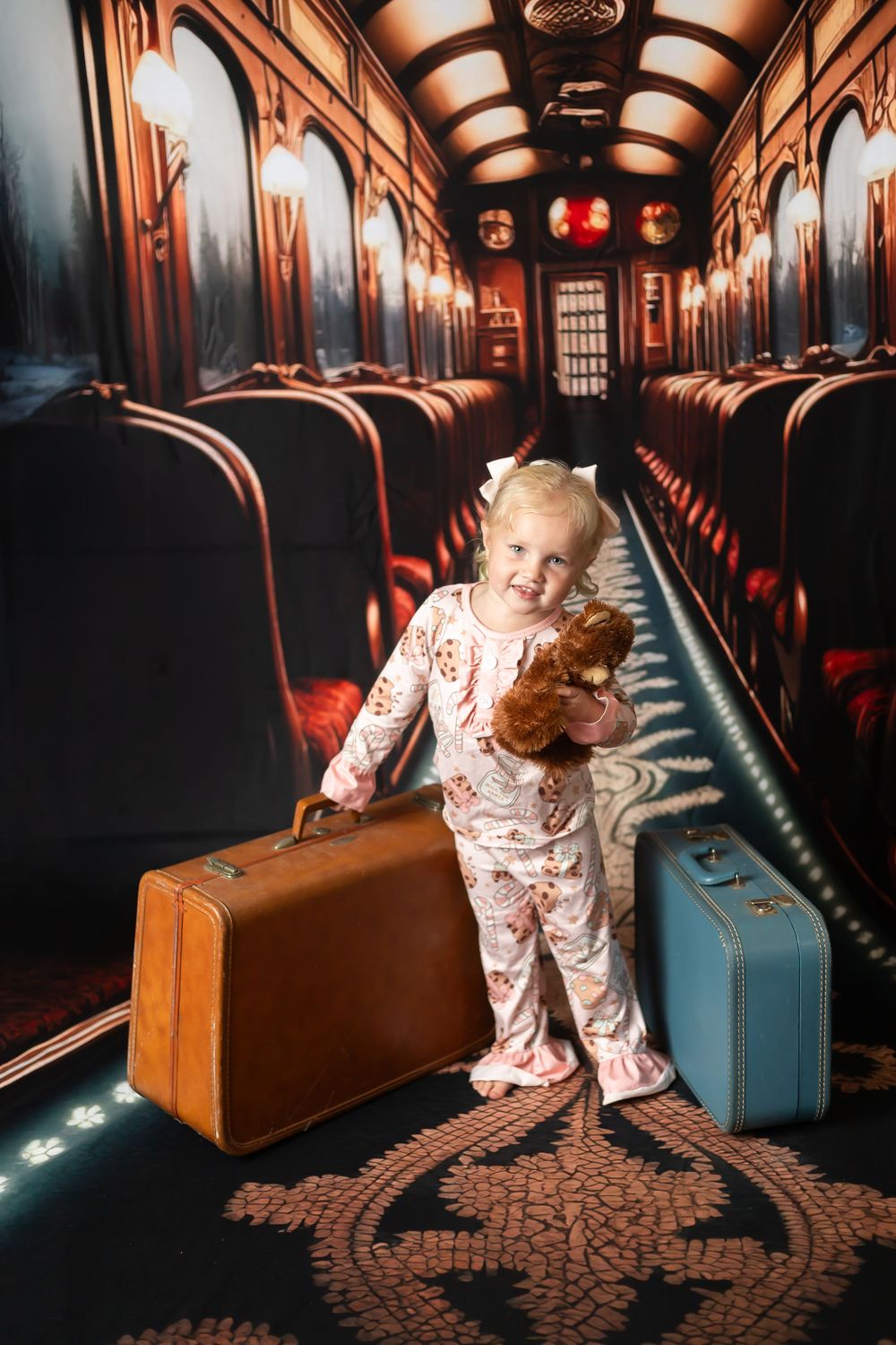 Toddler girl in pajamas holding a teddy bear and vintage suitcase in a decorated train corridor backdrop.