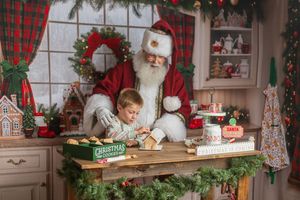 A young boy sits with Santa Claus at a festive wooden table decorated with Christmas greenery, making holiday cookies together.