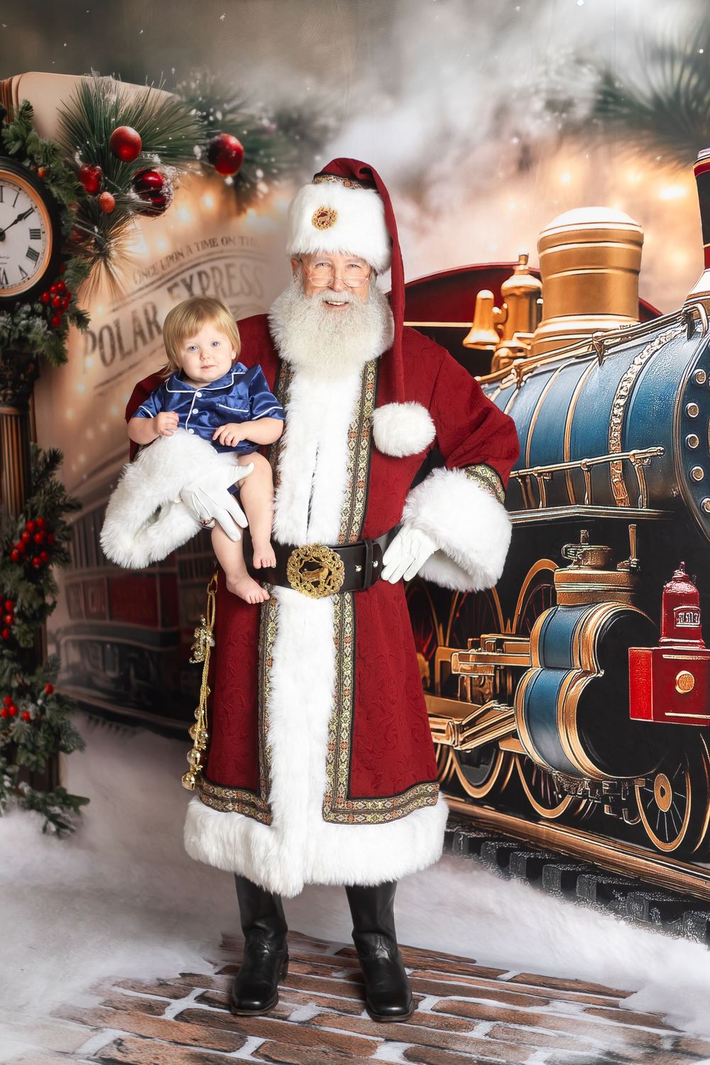 Santa Claus holds a baby in front of a Polar Express train backdrop with festive holiday decorations.