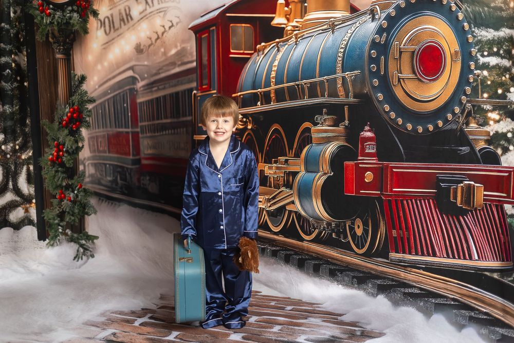 Boy in blue pajamas holding a suitcase poses before a Polar Express train mural at a Christmas photo set.