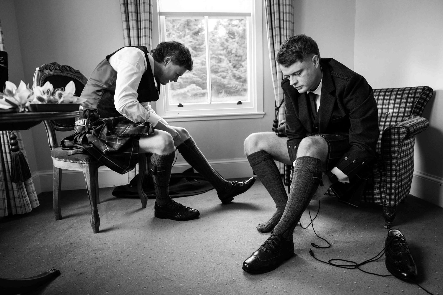 Two groomsmen in kilts putting on tall dress socks and shoes before a Scottish wedding ceremony.
