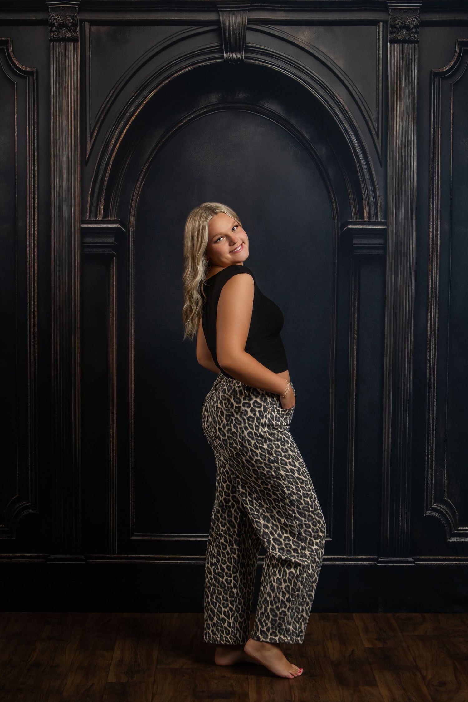 Pregnant blonde woman in black top and leopard print pants poses barefoot against dark ornate wall.