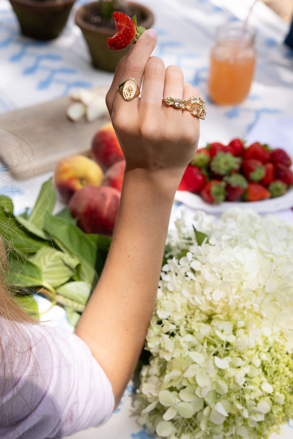 A woman wearing gold rings holds a fresh strawberry above a table with peaches, strawberries, and white hydrangeas.
