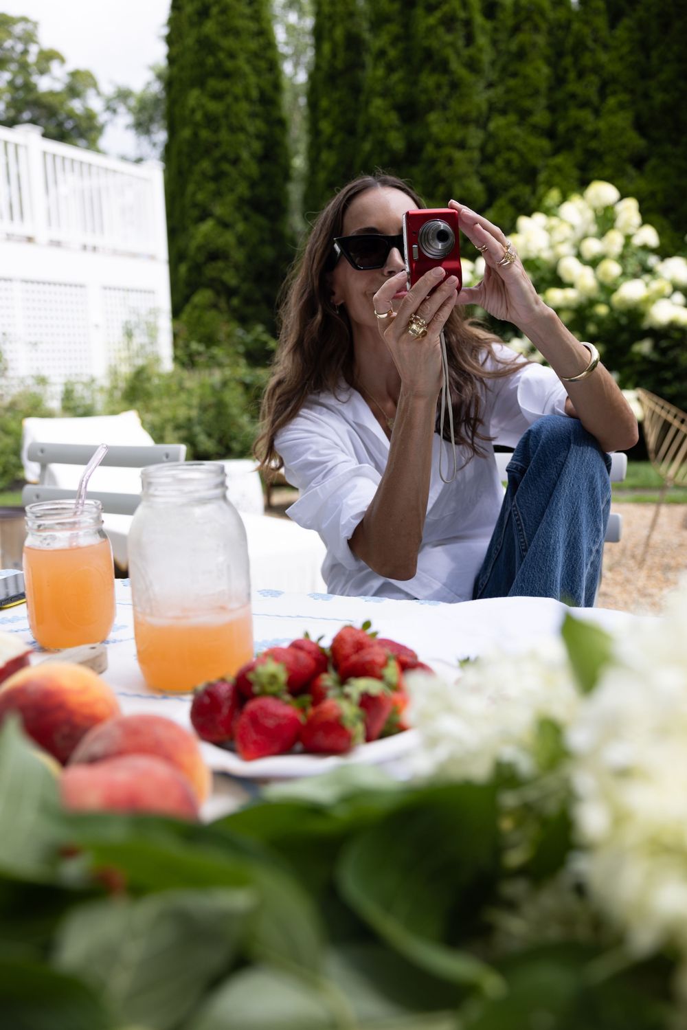 A stylish woman in sunglasses photographs a summer table spread with fruit, mason jar drinks, and hydrangeas outdoors.