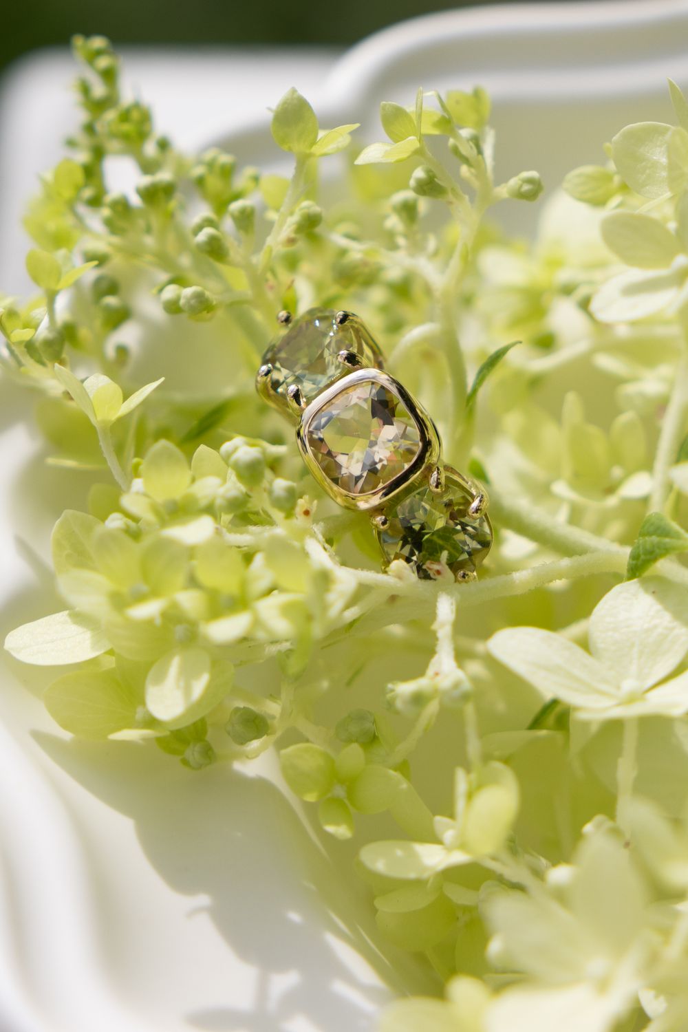 Gold ring with a cushion-cut green gemstone nestled among white hydrangea blooms outdoors.