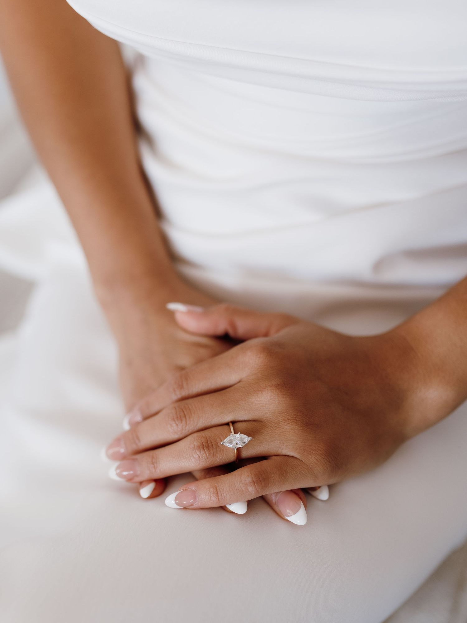 Close-up of a bride's hands with French manicure nails wearing an elegant marquise diamond engagement ring in gold setting.