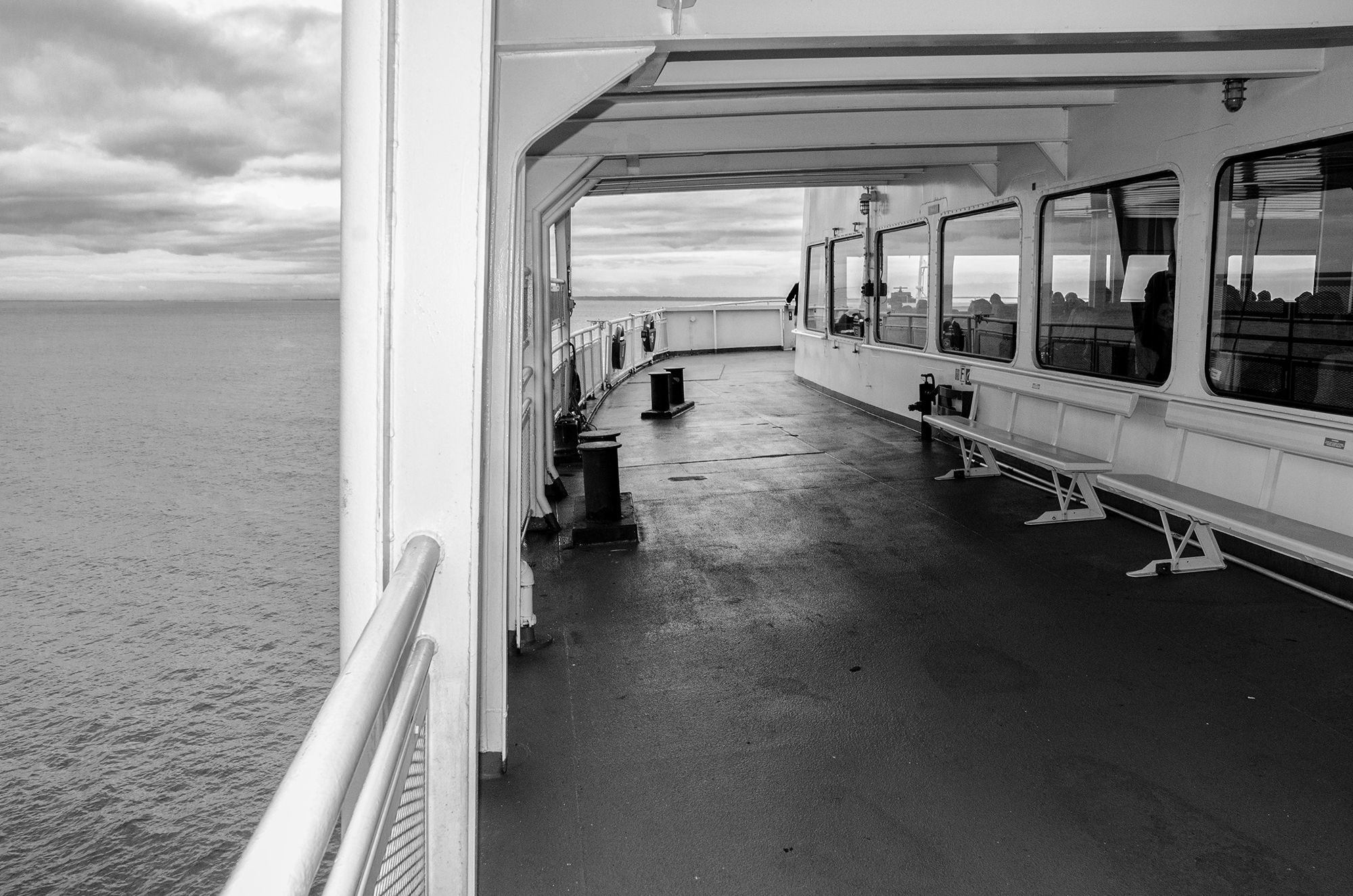 Black and white photo of an empty ferry deck with benches, railing, and calm ocean views under a cloudy sky.