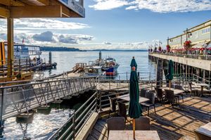 Waterfront wooden deck with outdoor café tables and closed green umbrellas overlooking calm blue water, boats, and a pier on a sunny day.