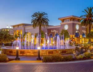 Illuminated fountain with blue lights at a shopping center plaza at dusk, framed by palm trees and a clock tower.