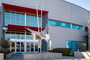 Abstract metal bird sculpture on a concrete pedestal outside a modern building with red awnings, glass windows, and gray corrugated siding.