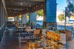 Covered outdoor restaurant patio with wooden tables and chairs, string lights, stone columns, and planter boxes on a sunny day.