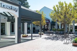 LaserAway storefront entrance beside a sunny brick-paved outdoor plaza with shaded patio tables, chairs, umbrellas and leafy trees.