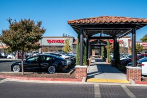 Black sedan and other cars parked beside a brick-pillared covered walkway leading to a T.J. Maxx storefront in a sunny shopping center.