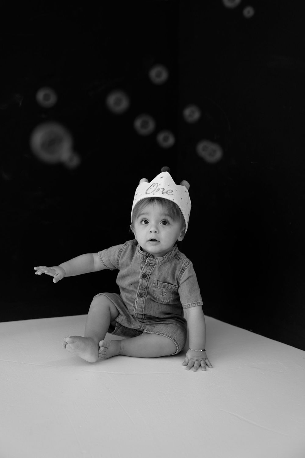 Black and white photo of a baby wearing a crown hat sitting on white floor, reaching out with one hand.