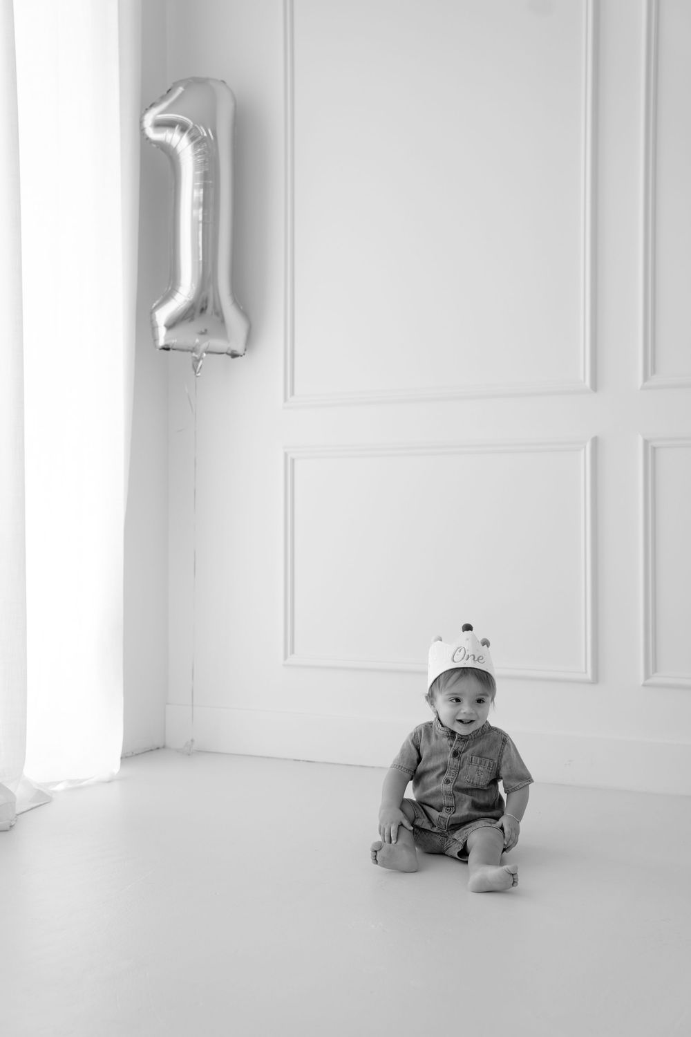 Black and white photo of a baby sitting on floor wearing a party hat next to a number one balloon for first birthday.
