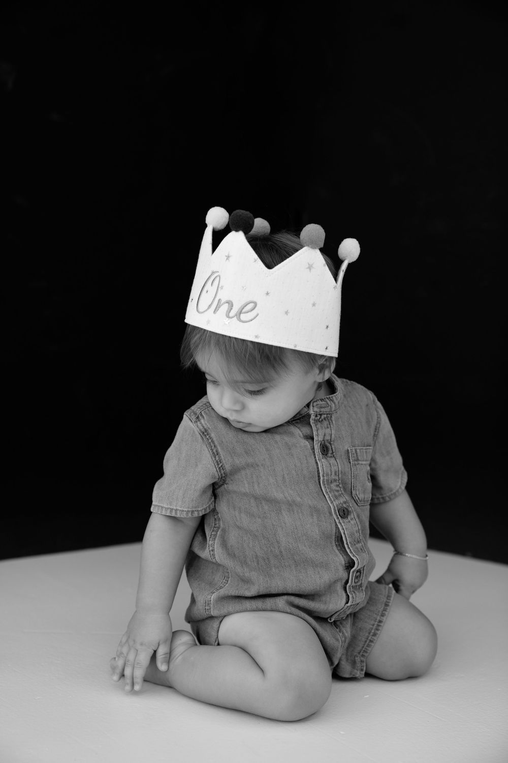 Black and white photo of a baby wearing a crown hat that says One, sitting cross-legged, celebrating their first birthday.