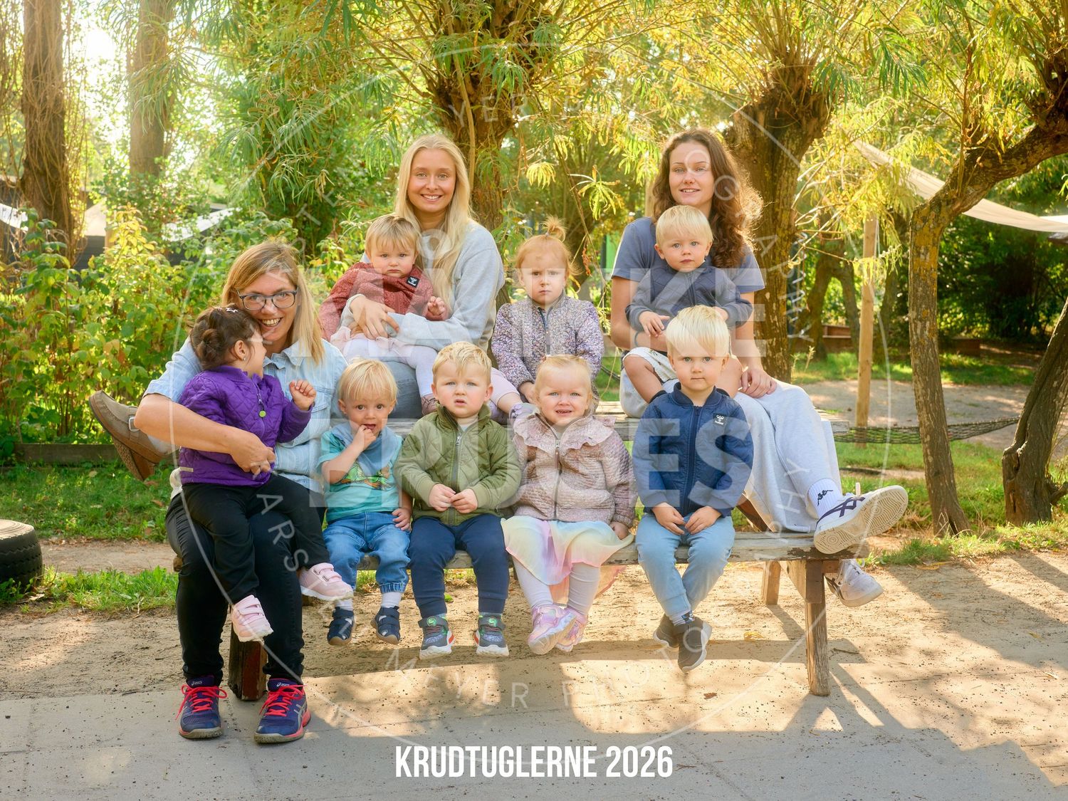 Group of women and toddlers posing outdoors on a bench surrounded by green trees, labeled Krudtuglerne 2026.