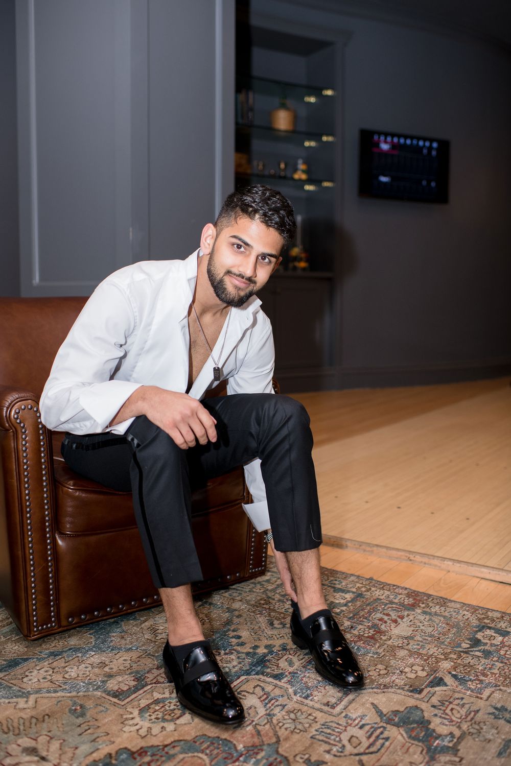 A stylish young man with a beard sits in a leather chair wearing a white shirt and black trousers with loafers.