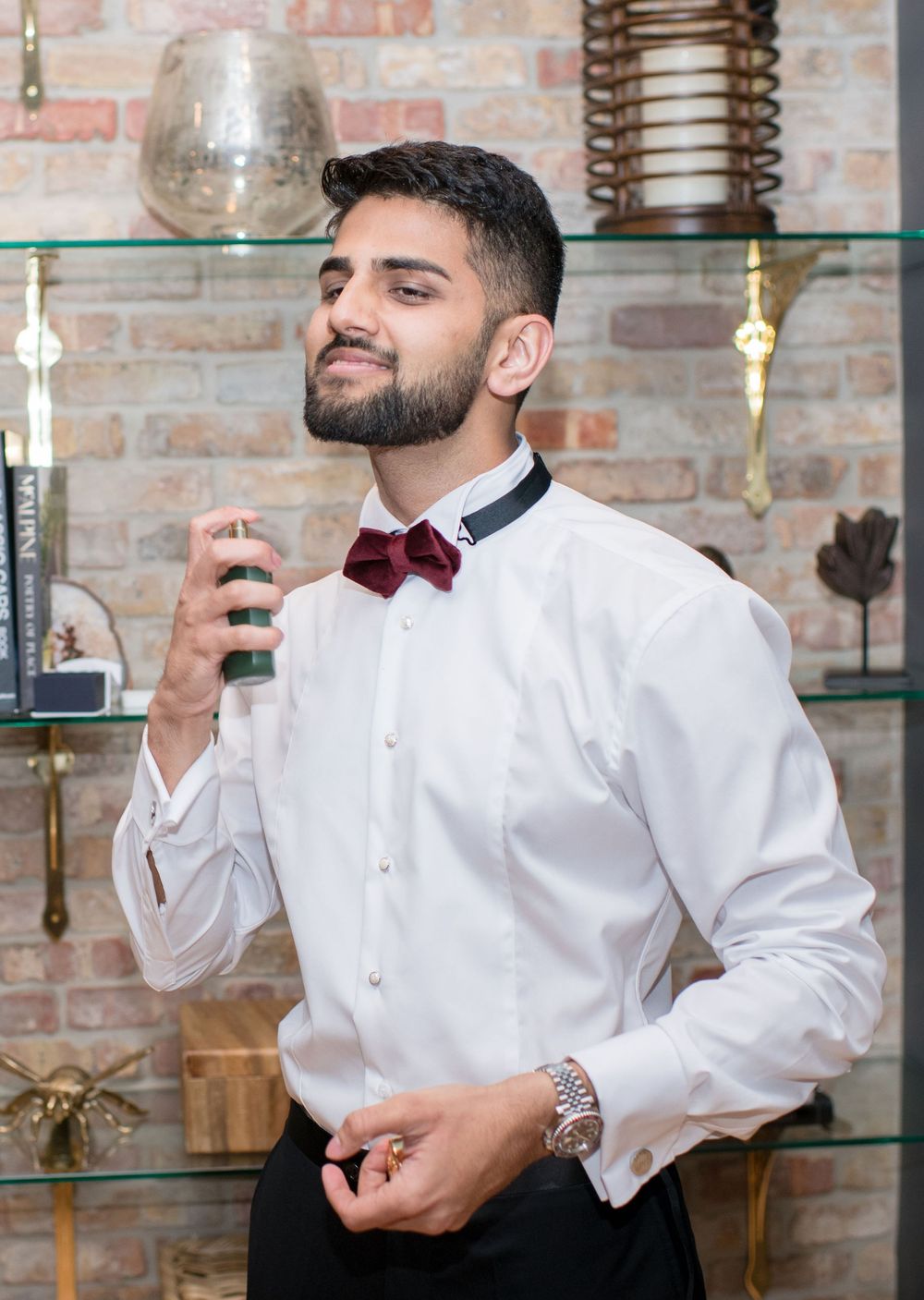 Stylish man in white dress shirt and burgundy bow tie spraying cologne, standing near brick wall with decorative shelves.