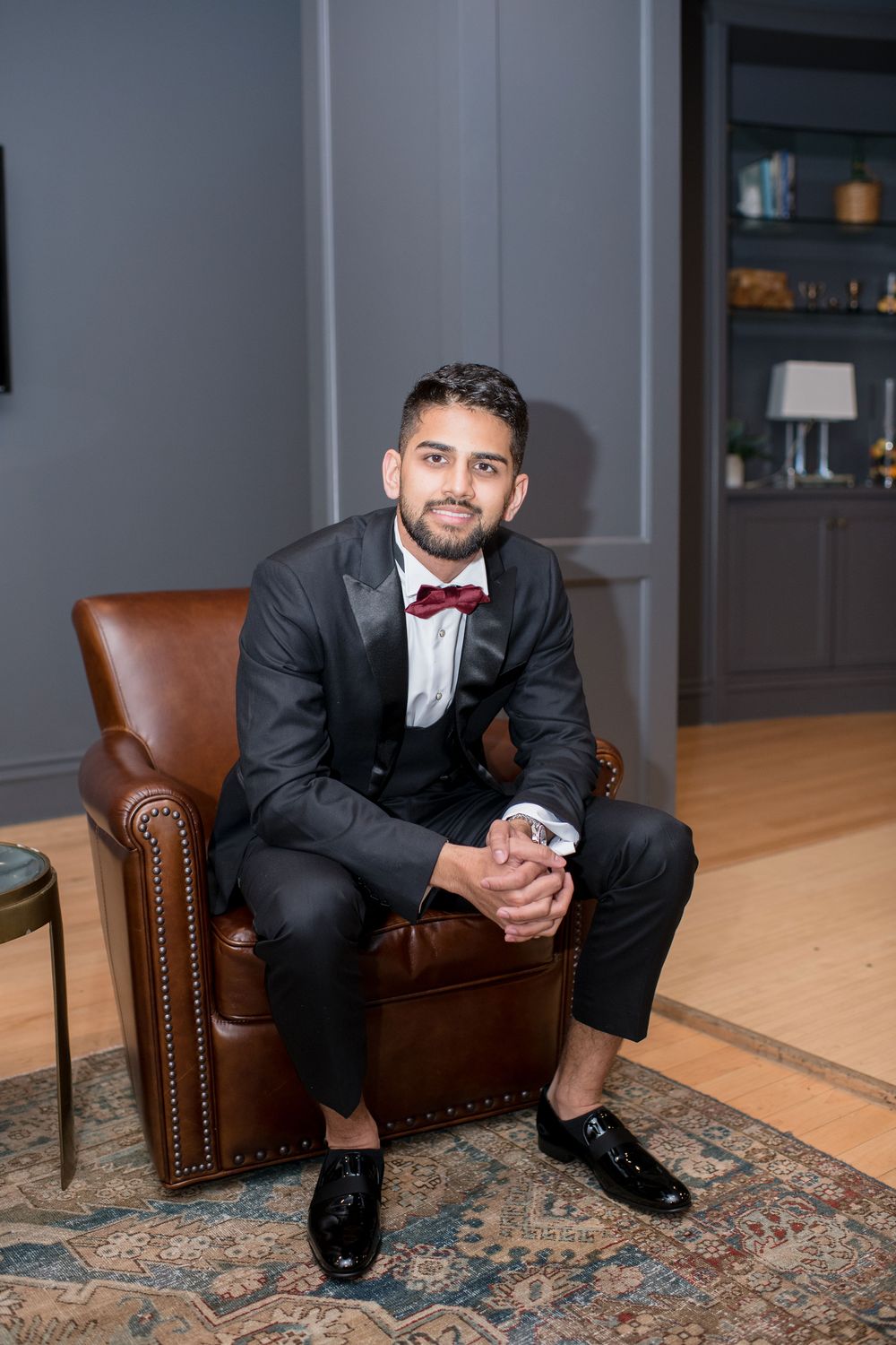 Dapper groom in black tuxedo with burgundy bow tie seated in leather armchair for formal portrait.