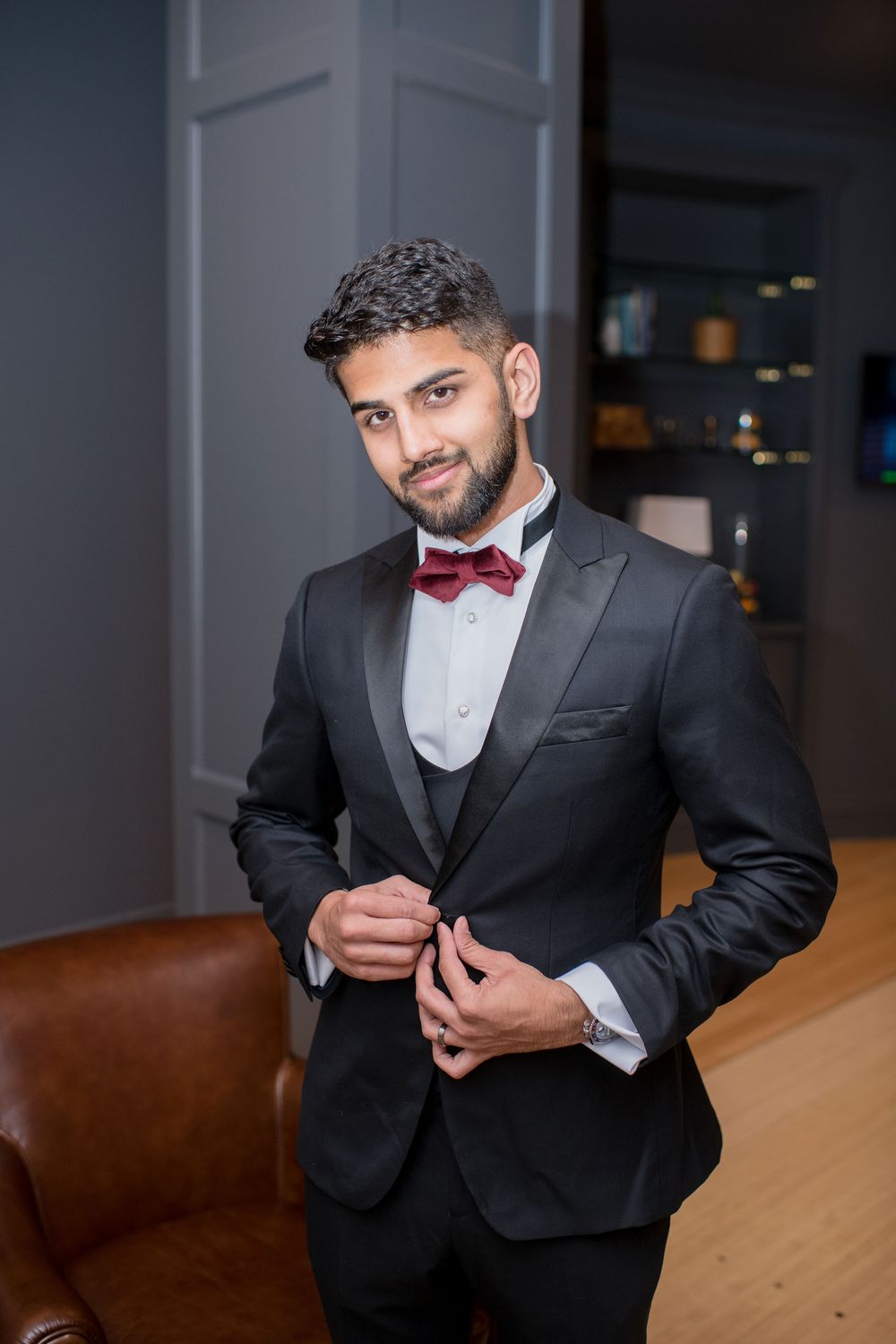 Elegant man in black tuxedo with burgundy bow tie buttoning jacket in stylish room.